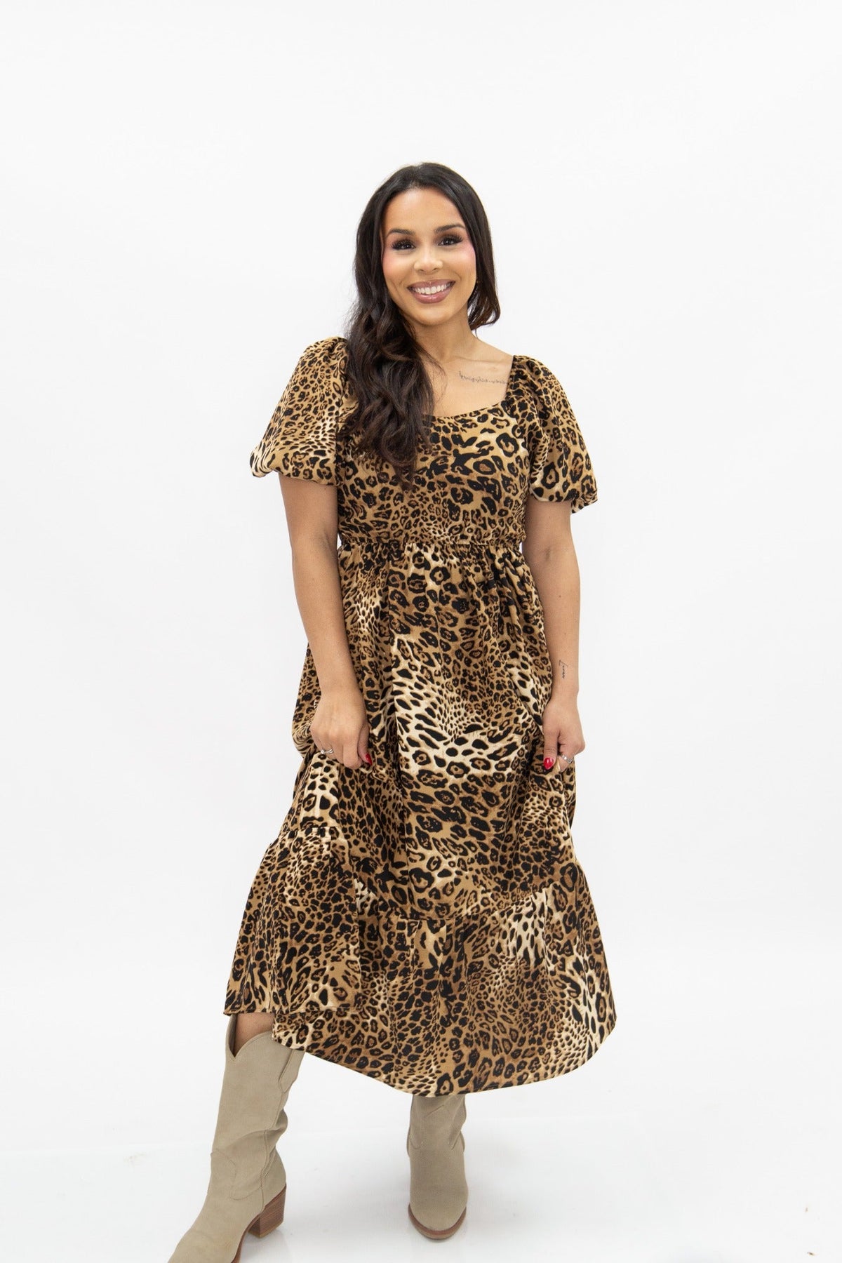 A smiling model wears a leopard print midi dress with puff sleeves and beige boots against a white background.