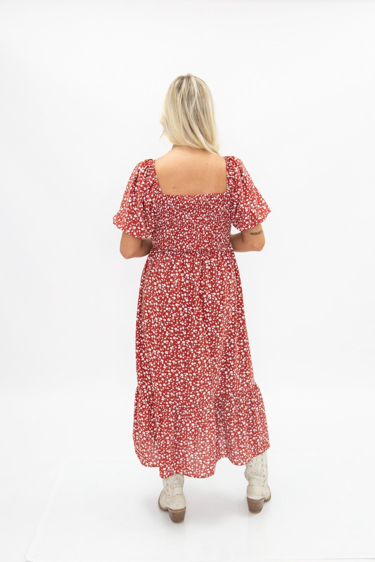 Back view of a blonde woman wearing a red floral puff sleeve midi dress with a square neckline and white boots against a white backdrop.