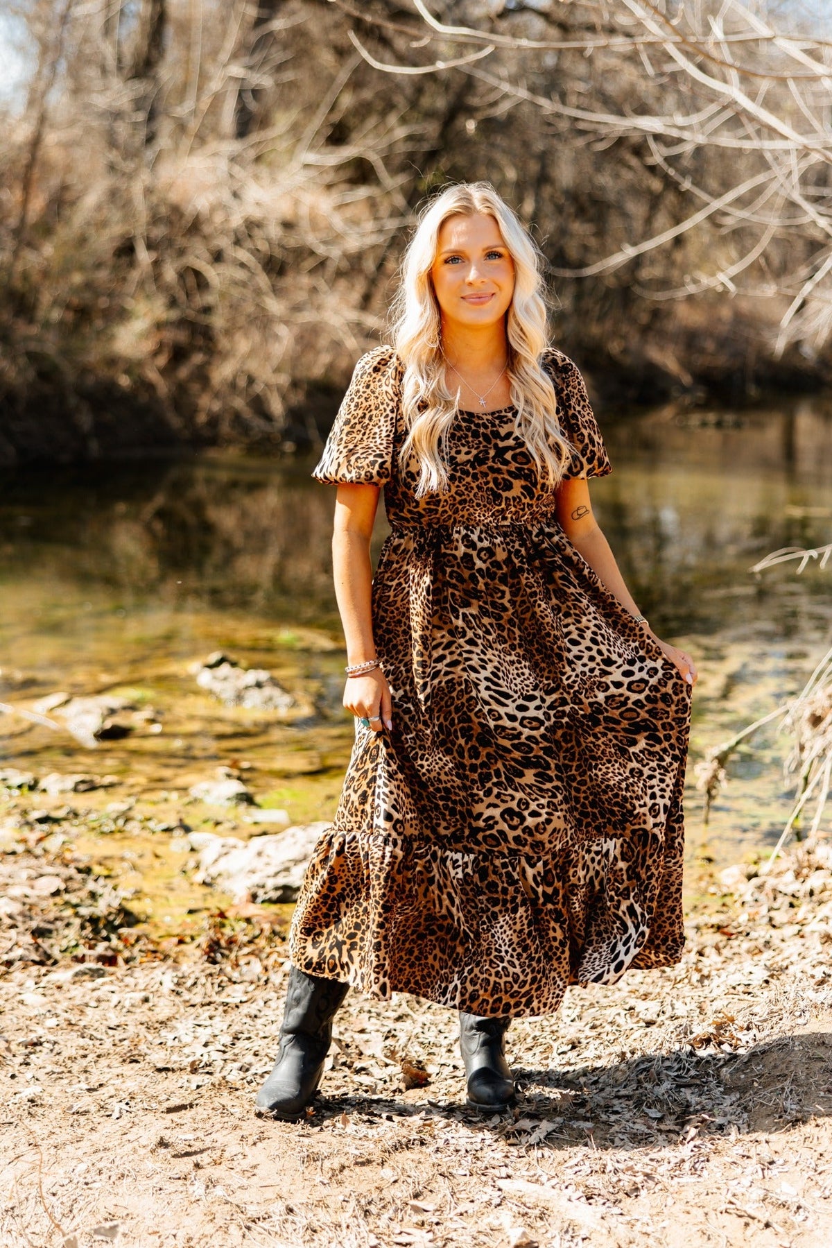 Blonde woman wearing a leopard-print puff-sleeve midi dress and black boots by a stream outdoors.
