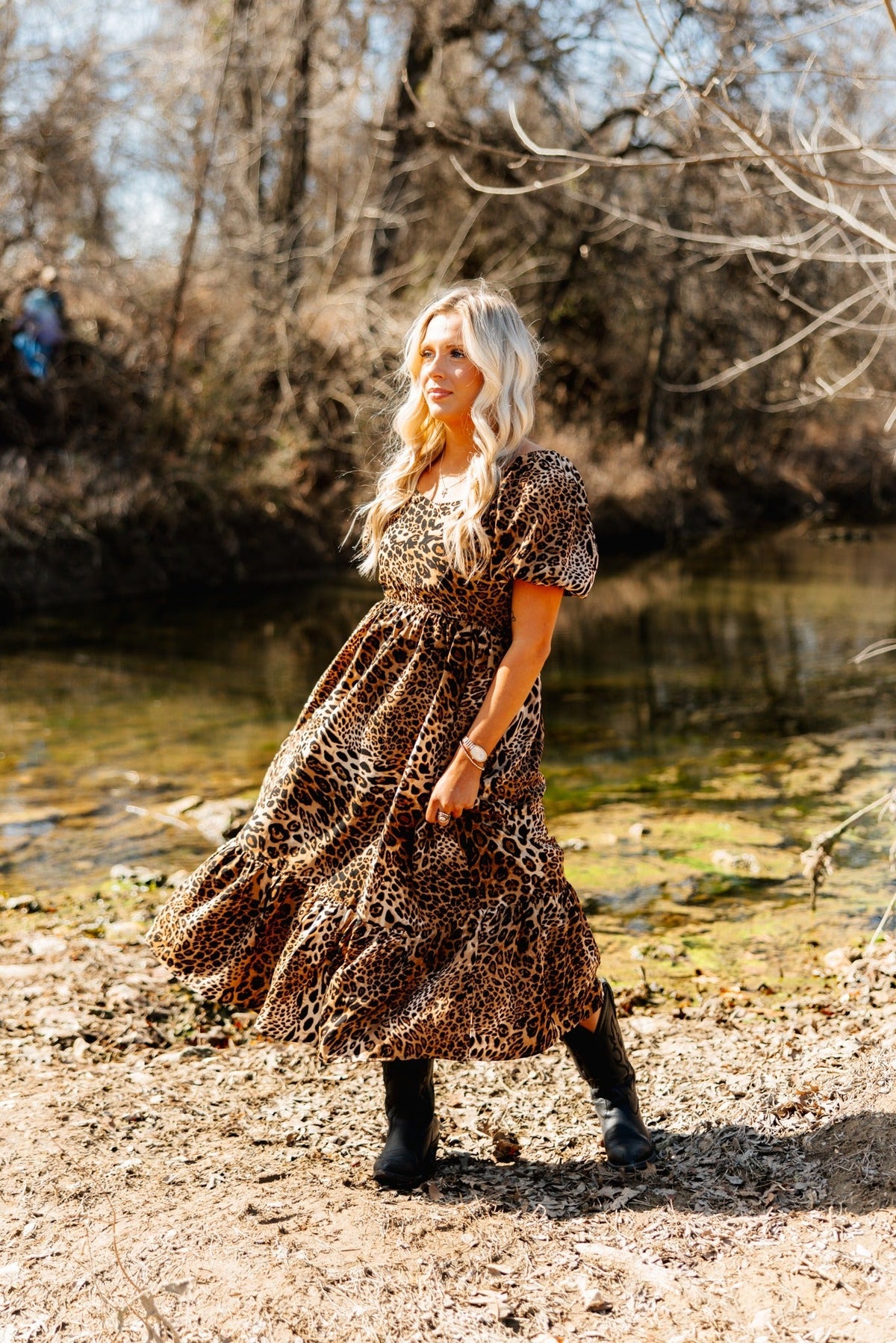 Blonde woman in a leopard print puff-sleeve midi dress and black boots by a stream.