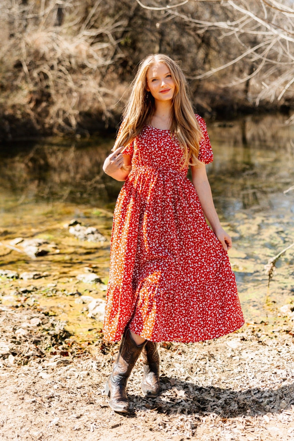 Blonde woman wears a red floral midi dress with puff sleeves and cowboy boots, standing by a stream.
