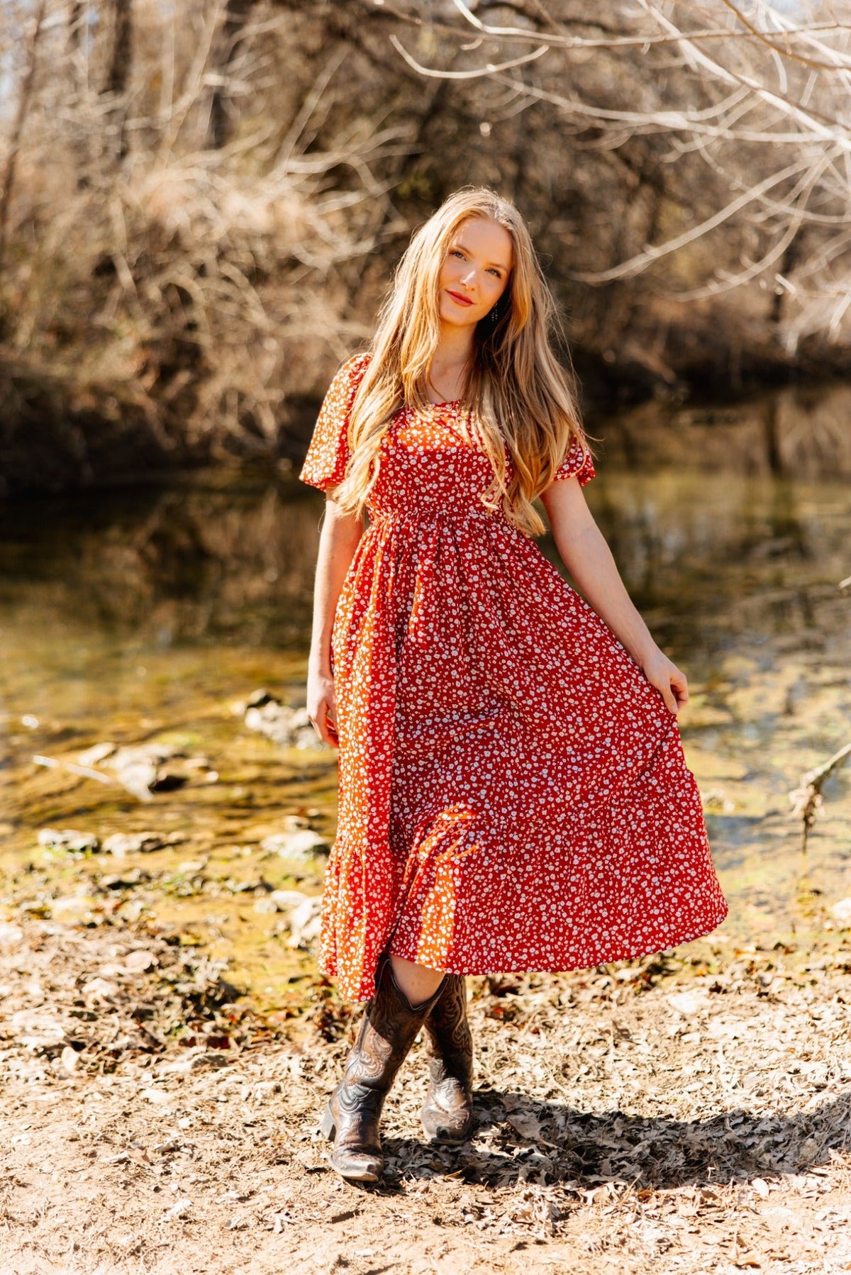 Blonde woman posing outdoors by a riverbank in a red floral puff-sleeve midi dress and cowboy boots.