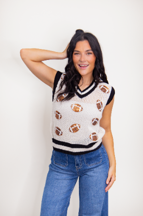 A woman models a white knit V-neck vest featuring sequined football patches and black striped trim, paired with blue jeans.