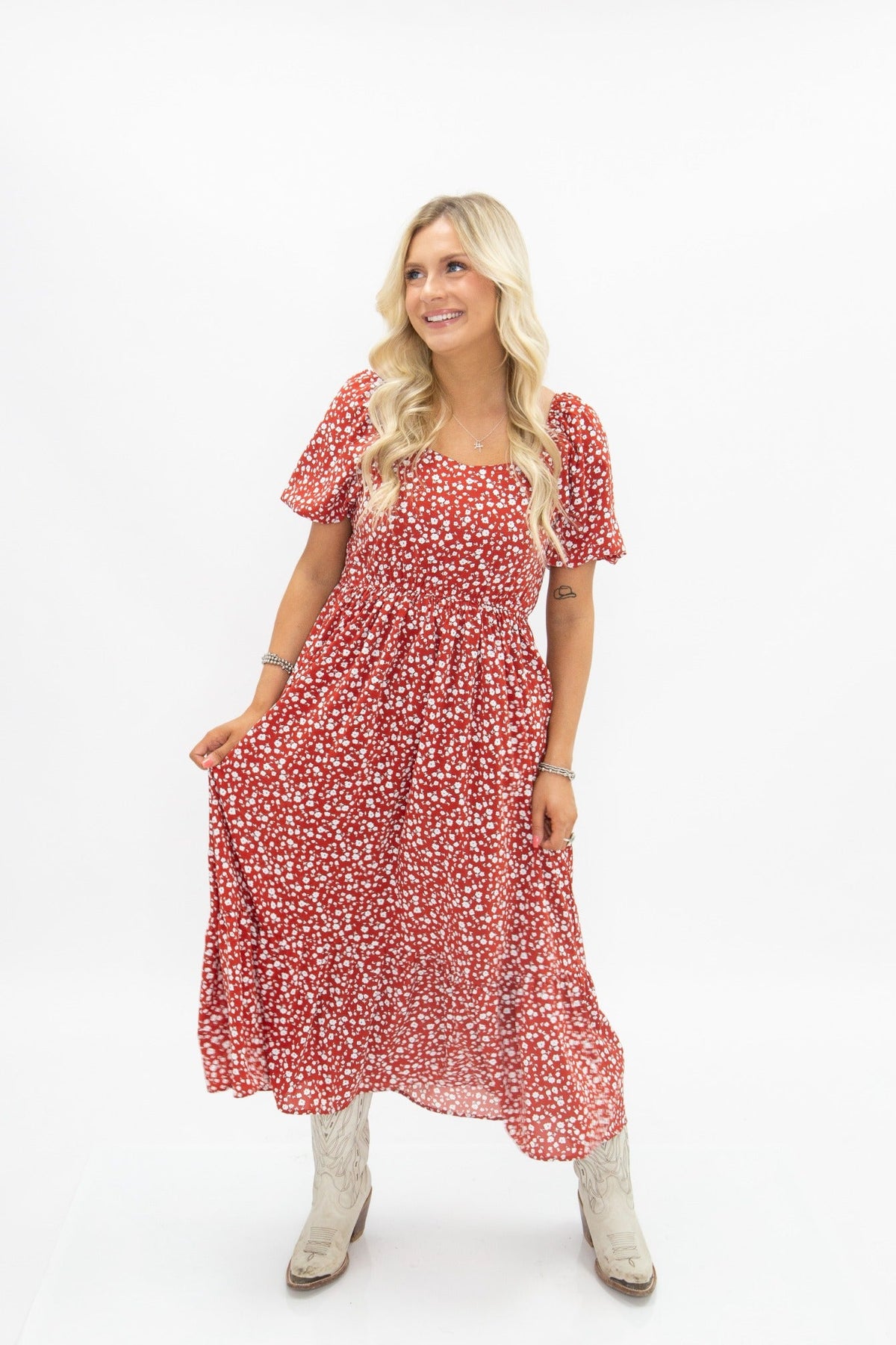 Blonde model poses in a red puff-sleeve midi dress with a white floral print, wearing white cowboy boots, against a white background.